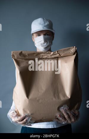 Safe food or goods delivery. Young courier delivering grocery brown eco paper bag order to the home of customer with mask and gloves during the corona Stock Photo