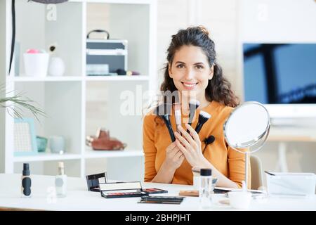Portrait of attractive young adult woman sitting at table holding various make-up brushes, looking at camera smiling Stock Photo