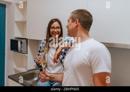 Beautiful young caucasian woman and handsome man chatting and drinking wine in the kitchen Stock Photo