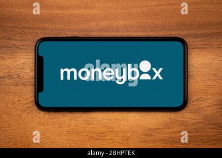 A smartphone showing the Moneybox logo rests on a plain wooden table (Editorial use only). Stock Photo