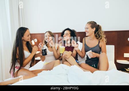 A group of women sitting on a bed eating from tubs of ice cream and smiling. Stock Photo