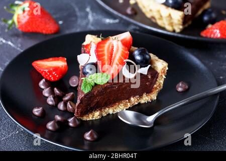 Piece chocolate tart with fresh strawberries, blueberries, coconut chips on top, the no-bake crust of rolled oats, dates and nuts served on a black pl Stock Photo