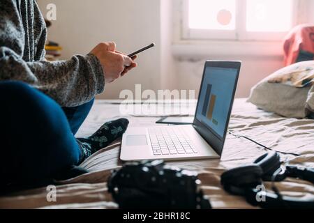 Close up of person sitting on bed, working on mobile phone and laptop computer during Corona virus crisis. Stock Photo