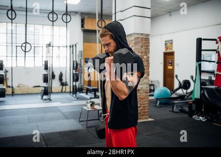 A man standing in a gym lifting a large dumbell with his left arm. Stock Photo