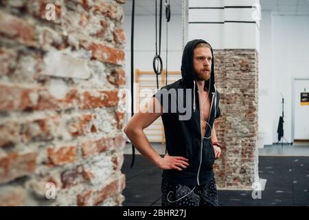 A man wearing a black hoodie with earphones in his ears standing in a gym with hands on his hips. Stock Photo