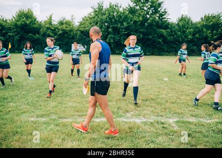 Group of women wearing blue, white and green rugby shirts on a training pitch, one running with a rugby ball with the coach watching. Stock Photo