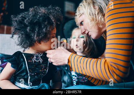 A woman bending down painting a child's face with another child watching in the background. Stock Photo