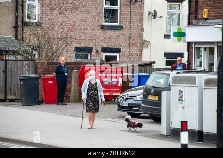 Sheffield UK –  March 30 2020: Elderly woman in PPE mask walks past socially distanced queue at Well Pharmacy during covid-19 pandemic, Richmond Road Stock Photo