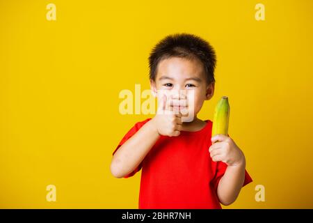 Happy portrait Asian child or kid cute little boy attractive smile wearing red t-shirt playing holds bananas and show finger thumb for good sign, stud Stock Photo