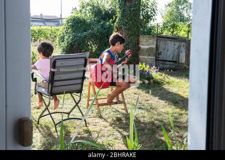 Two boys sitting on chairs in the garden, one playing a guitar. Stock Photo