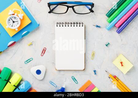 School stationery lies on a grey textured background with a Notepad in the center. The color of the rainbow, pencils, glasses, Notepad and Alarm clock Stock Photo