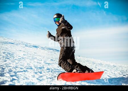 A person wearing a black ski suit, helmet and goggles skiing down a mountain on a red snowboard. Stock Photo