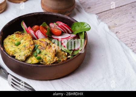 Zucchini fritters, vegetarian dish with freshly grated zucchini, egg yolk, pistachios and spring onion in rustic ceramic bowl served with green salad, Stock Photo
