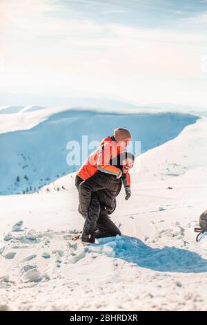 A man wearing a ski suit kneeling in the snow and giving a woman a piggy back, laughing and looking at the camera. Stock Photo