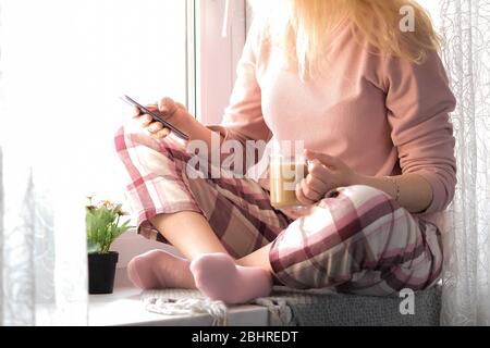 Cropped image of a blonde woman in her pijamas sitting on a window sill with a cup of coffee and her smartphone in hands. Self isolation, stay home or quarantine concept. Stock Photo