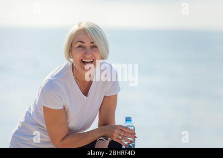 Portrait of a mature smiling woman after jogging. Senior lady drinking water  on background the sea and blue sky Stock Photo