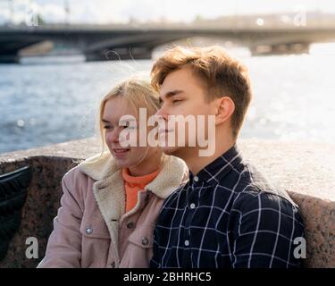 Teenagers in love sit on the waterfront in the city center, resting in autumn sun. Concept the first teenage love, the beginning of relations. A boy Stock Photo