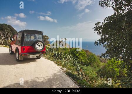 A bright red jeep on a road on the edge of a cliff overlooking the sea Stock Photo