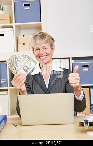 Elderly smiling businesswoman in the office with lots of dollar bills holds thumbs up Stock Photo