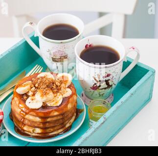 Stack of pancakes with maple syrup, chopped bananas and pecan nuts. Two cups of coffee on breakfast tray. Stock Photo