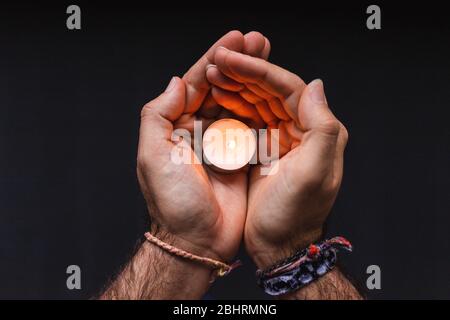 two hands with bracelets holding a small burning candle on black background Stock Photo