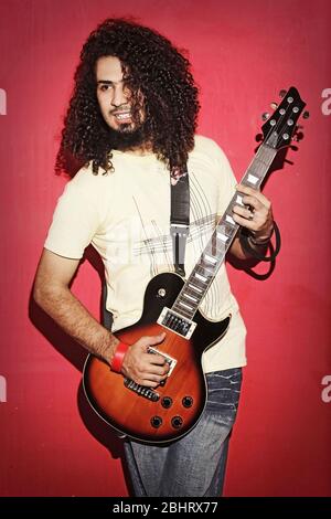 Closeup of one handsome passionate expressive cool young brunette rock musician men with long curly hair playing electro guitar standing against red b Stock Photo