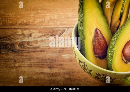 Close-up photo of fresh sliced avocados, brown seeds visible in green bowl on brown wooden background. Vegetarian food concept, vintage style. Stock Photo