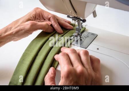 Close-up view on the hands of a woman using a sewing machine to sew homemade reusable cloth face masks in green cotton fabric. Stock Photo