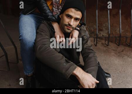Handsome man with beard sitting on the road and woman hands on his shoulders Stock Photo