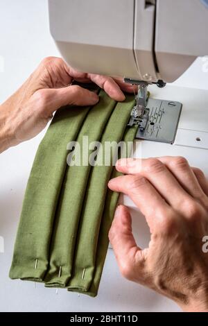 Close-up view on the hands of a woman using a sewing machine to sew homemade reusable cloth face masks in green cotton fabric. Stock Photo