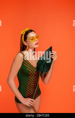 Woman in swimsuit and sunglasses looking at green leaf isolated on orange Stock Photo
