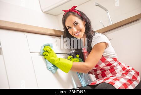 Lovely young housemaid in apron cleaning white kitchen cupboard door with a cloth and detergent in yellow rubber gloves. Stock Photo