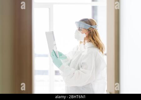 A female doctor using a tablet wearing a safety suit, protective mask and latex gloves. A blonde Caucasian woman protected from infection or pandemic Stock Photo