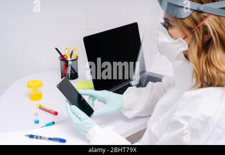 A female doctor uses the mobile phone at the laboratory desk. There are syringes and she is wearing a safety suit, protective mask and latex gloves. Stock Photo