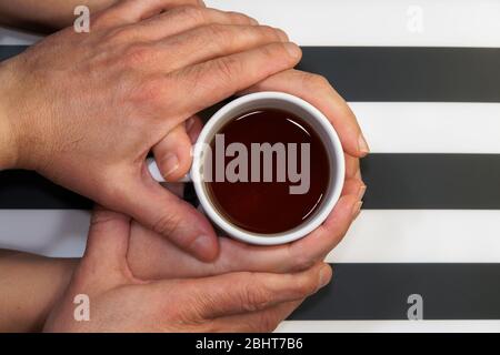 Loving couple on relationship, affectionately holding hands over beverage cup top view.Male tenderly touching female hands that hold a tea mug at home