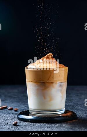 Iced Dalgona Coffee and Sifting Cocoa on Dark Background. Trendy Creamy Whipped Coffee. South Korean Cold Summer Drink. Stock Photo
