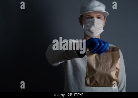 Safe food or goods delivery. Young courier delivering small brown eco paper bag order to the home of customer with mask and gloves during the coronavi Stock Photo