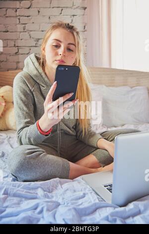 A blonde girl is sitting on the bed in a gray tracksuit with a phone and laptop.  Stock Photo