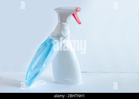 The bottle spray of white color with the red button and the medical mask of blue color is on a table of the white color, a set during a pandemic for Stock Photo