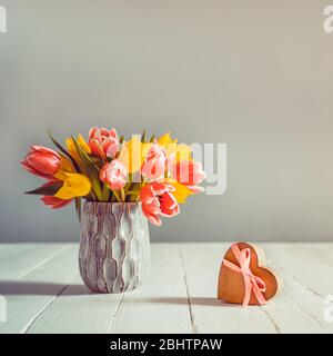 Bouquet of pink and yellow tulip flowers in a blue vase and heart shape gift box with satin ribbon bow on the white wooden table and grey background. Stock Photo