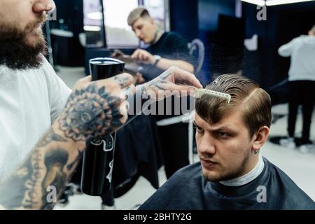 Attractive male is getting a modern haircut in barber shop. Tattooed Barber wets hair by spray and combs them. Stock Photo