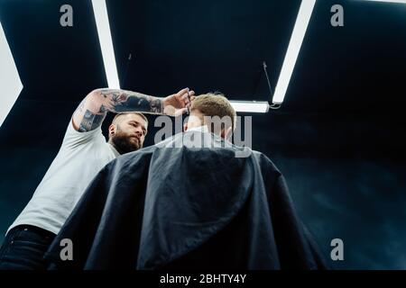 Tattooed Barber cuts the hair of the client with scissors. Close up. Attractive male is getting a modern haircut in barber shop. Bottom view. Stock Photo