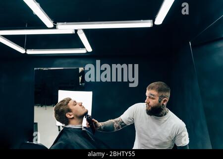 Tattooed Barber trimming bearded man with shaving machine in barbershop. Hairstyling process. Hairstylist cutting the beard of a bearded male. Stock Photo