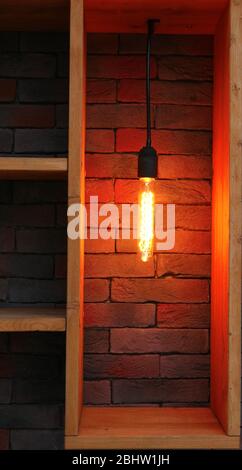 Vintage electric lamp hanging on the wooden shelving in front of brick wall. Warm light in retro design. Stock Photo