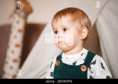 Sweet 1 year old little baby boy playing in a teepee game tent in a children's room decorated in pastel light colors, dressed in a green jumpsuit Stock Photo