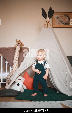 Sweet 1 year old little baby boy playing in a teepee game tent in a children's room decorated in pastel light colors, dressed in a green jumpsuit Stock Photo