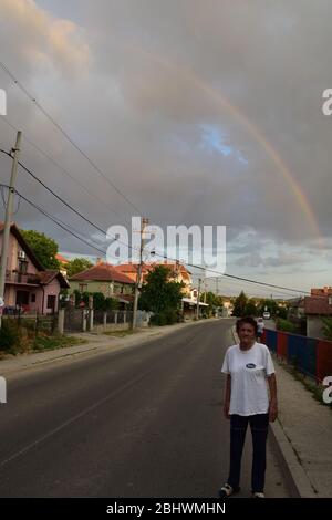 An elderly woman stands on the road by the sidewalk. Behind the woman, there is a rainbow in the sky Stock Photo