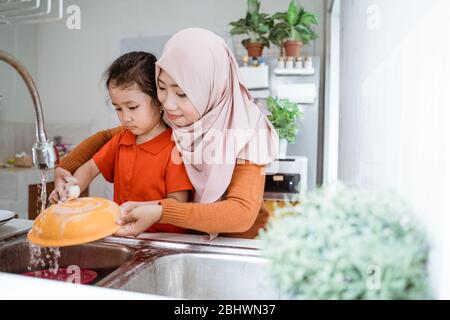 Helping hand. Cute little Girl Help Her muslim Mother In Washing Dishes At Family Kitchen Stock Photo