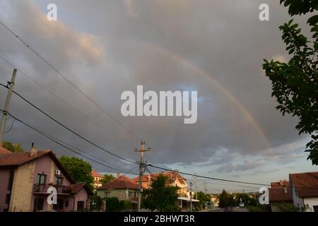 Houses in the village next to the road, and above them are gray clouds and rainbows across the clouds Stock Photo