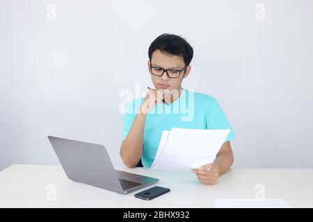 Young Asian Man is serious and focus when working on a laptop and document on the table. Indonesian man wearing blue shirt. Stock Photo